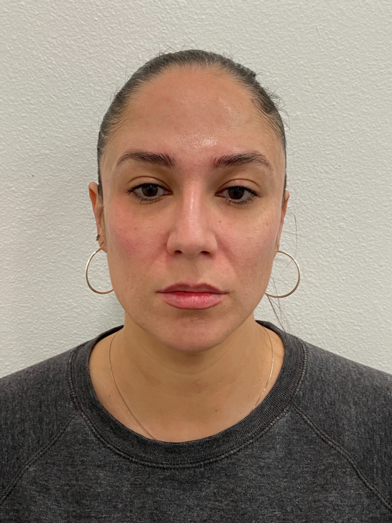 Portrait of a woman with long hair pulled back, wearing hoop earrings and a dark sweatshirt, against a white background. She has a neutral expression and minimal makeup, showcasing her natural features.