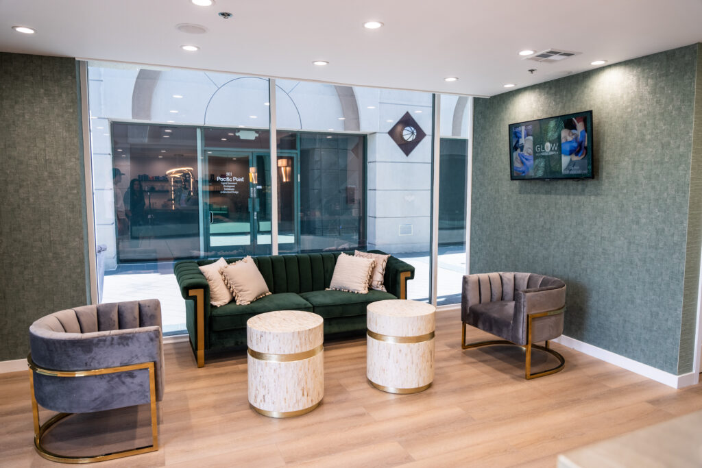 Modern waiting area featuring a green velvet sofa and two stylish circular tables, surrounded by gray accent chairs. Large windows provide natural light, showcasing an inviting atmosphere. Ideal for a spa or wellness center.