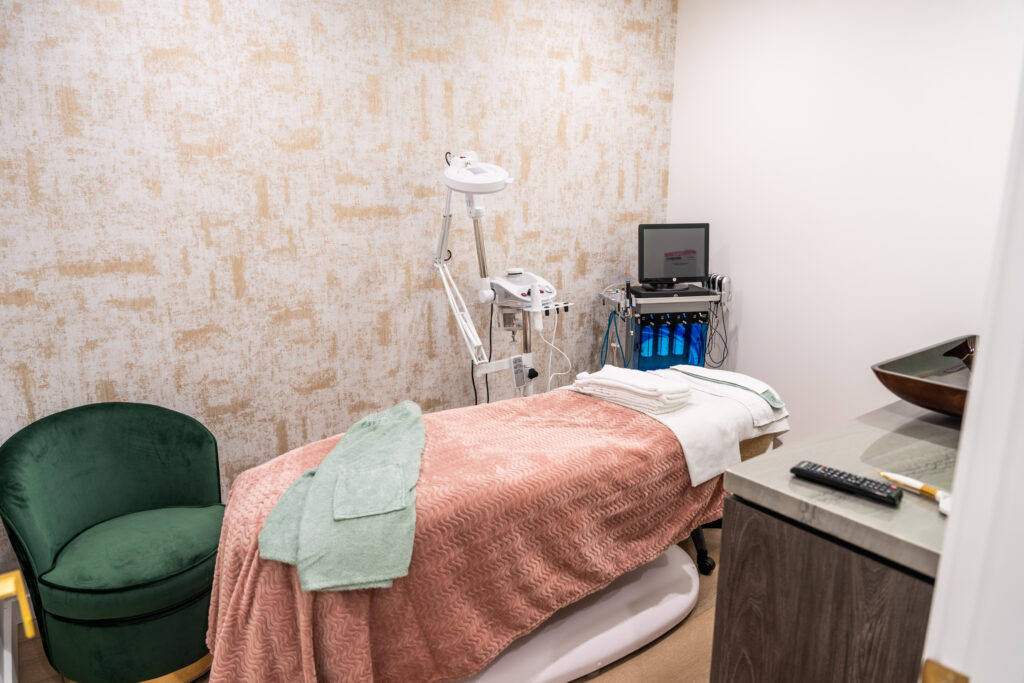 Spa treatment room featuring a massage bed with soft linens, a green armchair, and equipment for skincare treatments, including a monitor and various tools. The calming decor creates a relaxing atmosphere for clients.