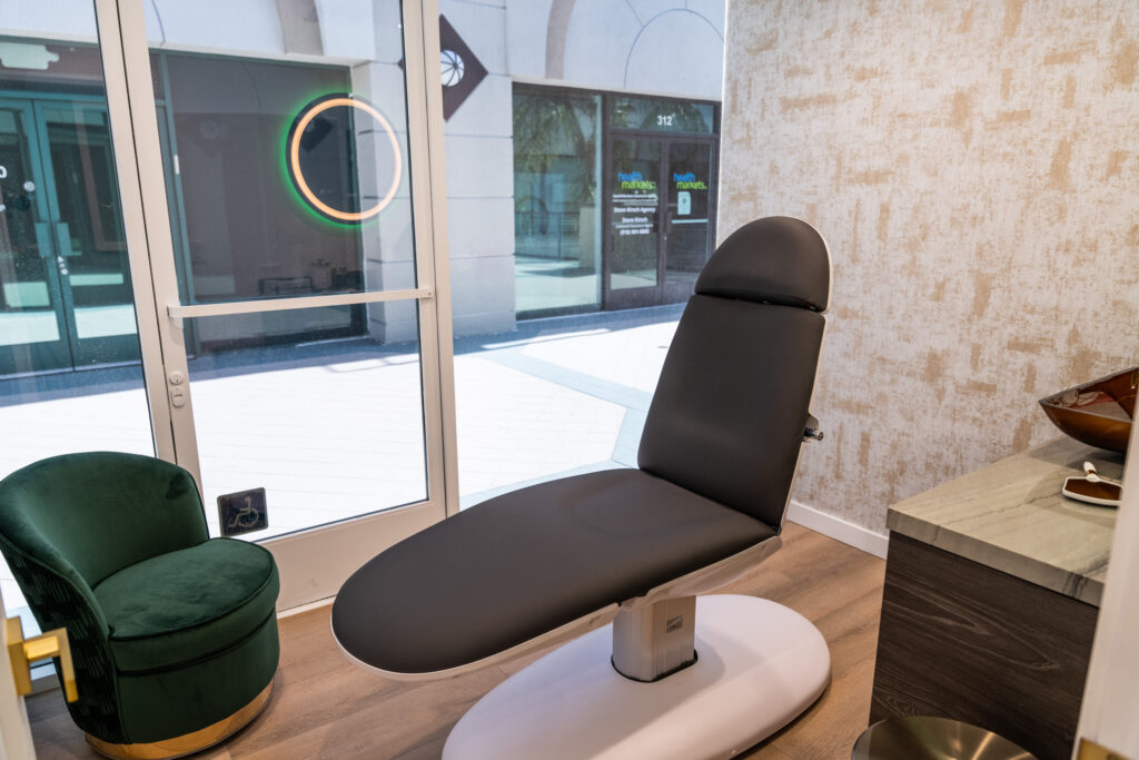 Interior view of a wellness treatment room featuring a modern treatment chair, a green velvet accent chair, and a stylish countertop. Natural light illuminates the space through a large window, enhancing the inviting atmosphere.