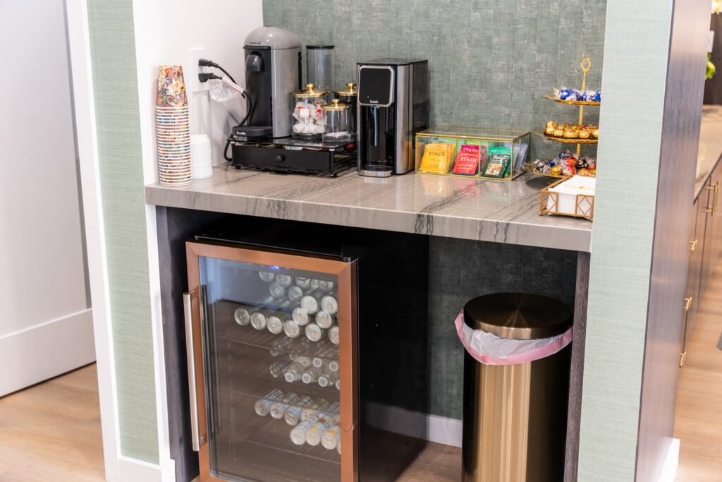 Coffee station setup featuring a coffee maker, tea selection, and a mini fridge stocked with drinks, alongside a decorative display of chocolates and a waste bin, all set against a stylish green wall.