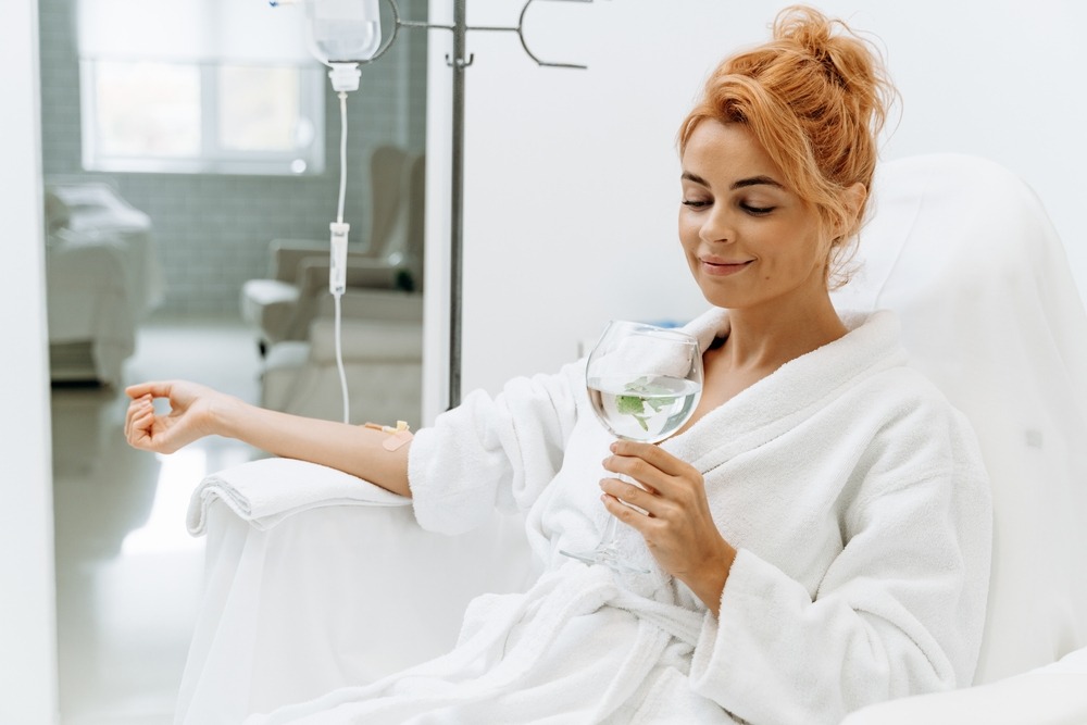 Woman in a white robe enjoying a glass of water with mint while receiving IV therapy in a bright, modern medical setting.